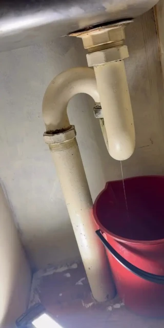 Water leaking from worn galvanised-steel drainage beneath a kitchen sink into a bucket